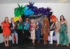 Group of people in colorful outfits wearing rainbow sashes posing with trophies at a Pride event, with a balloon arch and a black backdrop behind them.
