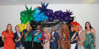 Group of people in colorful outfits wearing rainbow sashes posing with trophies at a Pride event, with a balloon arch and a black backdrop behind them.