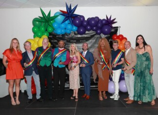 Group of people in colorful outfits wearing rainbow sashes posing with trophies at a Pride event, with a balloon arch and a black backdrop behind them.