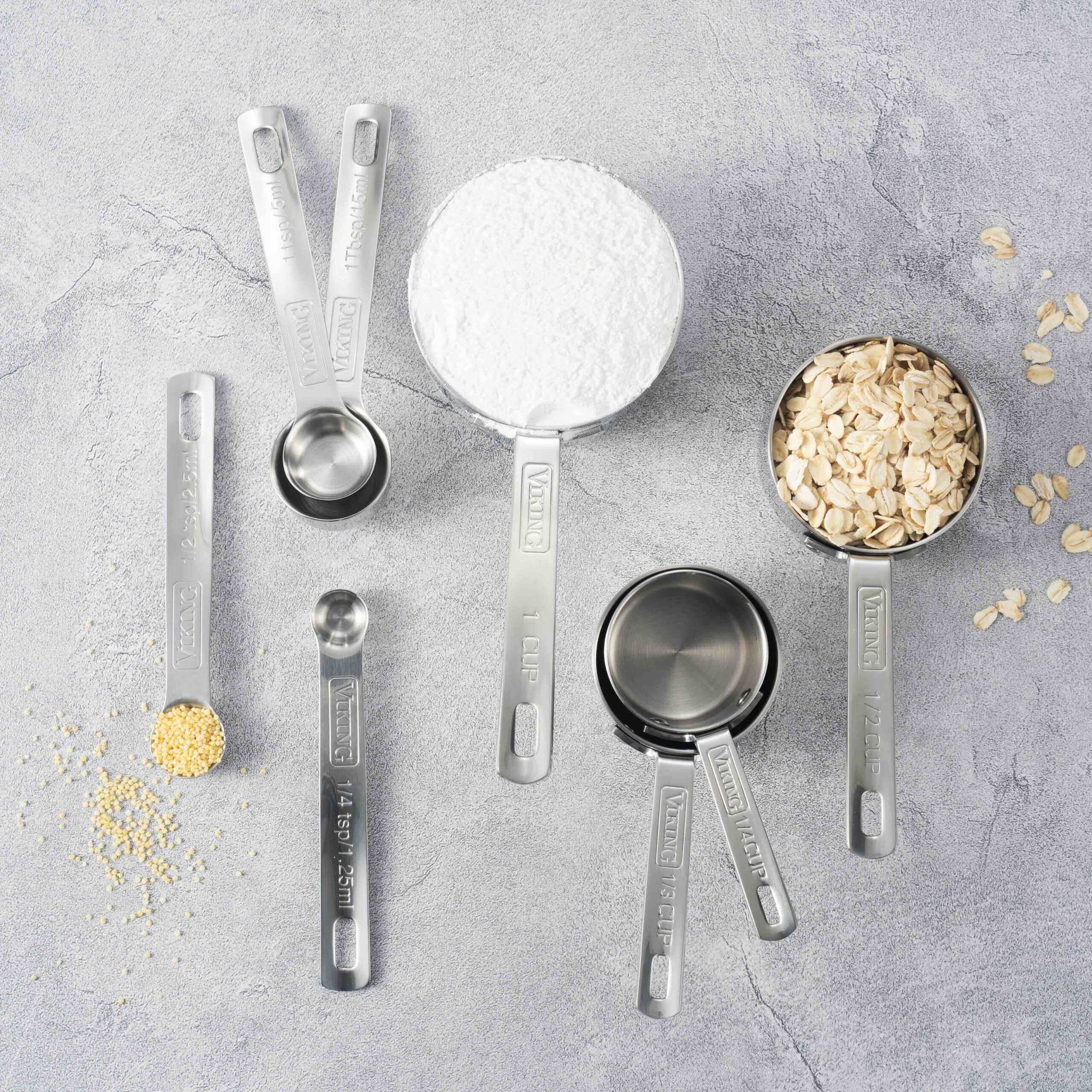 Top-down image of stainless measuring spoons and cups around bowls of flour and oats on a gray textured surface.