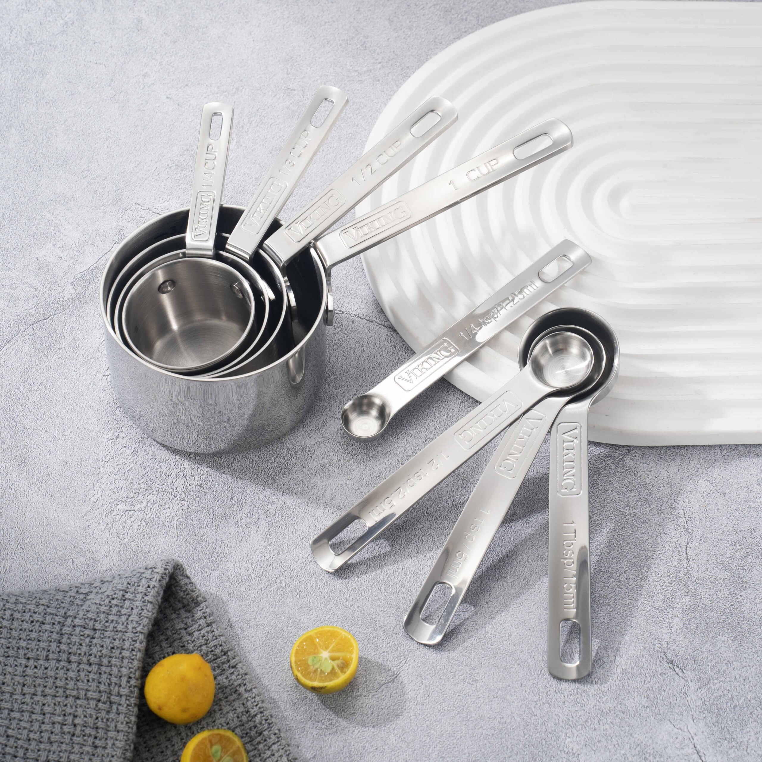 Top-down view of stainless steel measuring spoons spread around a white plate on a gray textured surface with lemon pieces nearby.”
