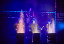 Four performers in blue-lit suits drum on stage, sparks flying from each drum as part of a dramatic percussion act.