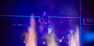 Four performers in blue-lit suits drum on stage, sparks flying from each drum as part of a dramatic percussion act.