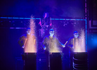 Blue Man Group – Comes to the Broward Center, June 11-14 Four performers in blue-lit suits drum on stage, sparks flying from each drum as part of a dramatic percussion act.