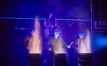 Four performers in blue-lit suits drum on stage, sparks flying from each drum as part of a dramatic percussion act.
