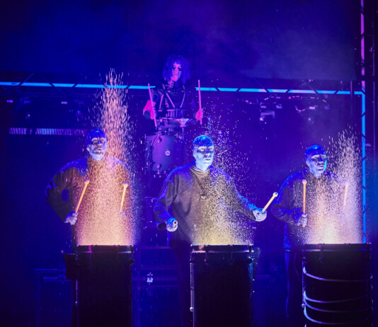 Four performers in blue-lit suits drum on stage, sparks flying from each drum as part of a dramatic percussion act.