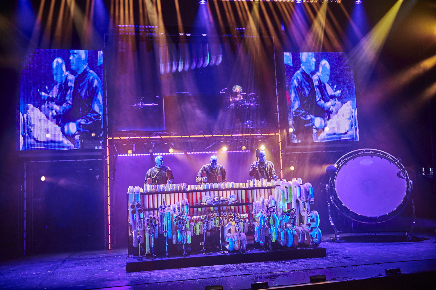 Live concert scene: three performers behind a long frame of colorful PVC pipes and bottles, under blue/purple lights, with a large drum on the right and video screens behind them.