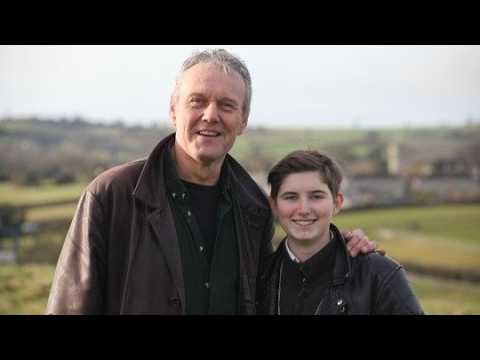 Two men posing outdoors with a rural landscape behind them, both smiling; older man on the left, younger man on the right.