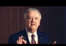 Older man in a suit gesturing with his hands while speaking in a formal setting.