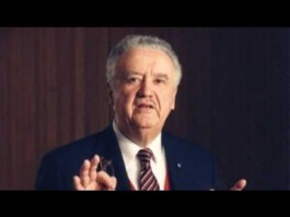 Older man in a suit gesturing with his hands while speaking in a formal setting.