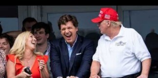 A group of people on a balcony laughing and chatting at a public event: a woman in a red dress, a man in a blue blazer leaning on the railing, and a man in a white polo with a red cap beside them.