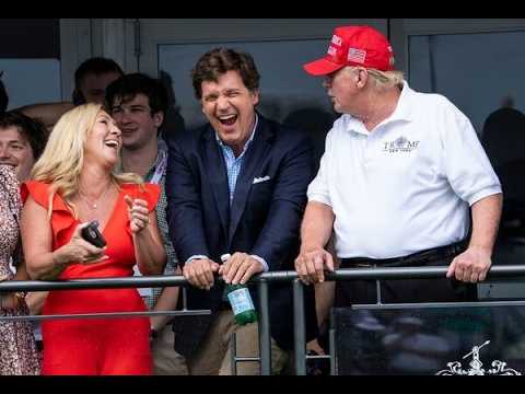 A group of people on a balcony laughing and chatting at a public event: a woman in a red dress, a man in a blue blazer leaning on the railing, and a man in a white polo with a red cap beside them.