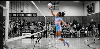 Girl volleyball player in a blue-pink uniform leaps to spike the ball over the net during a match at Central High, with a crowded gym in the background.