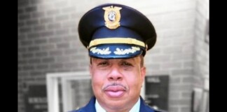Portrait of a man in a dark blue police uniform and peaked cap with gold insignia, outdoors in front of a building.