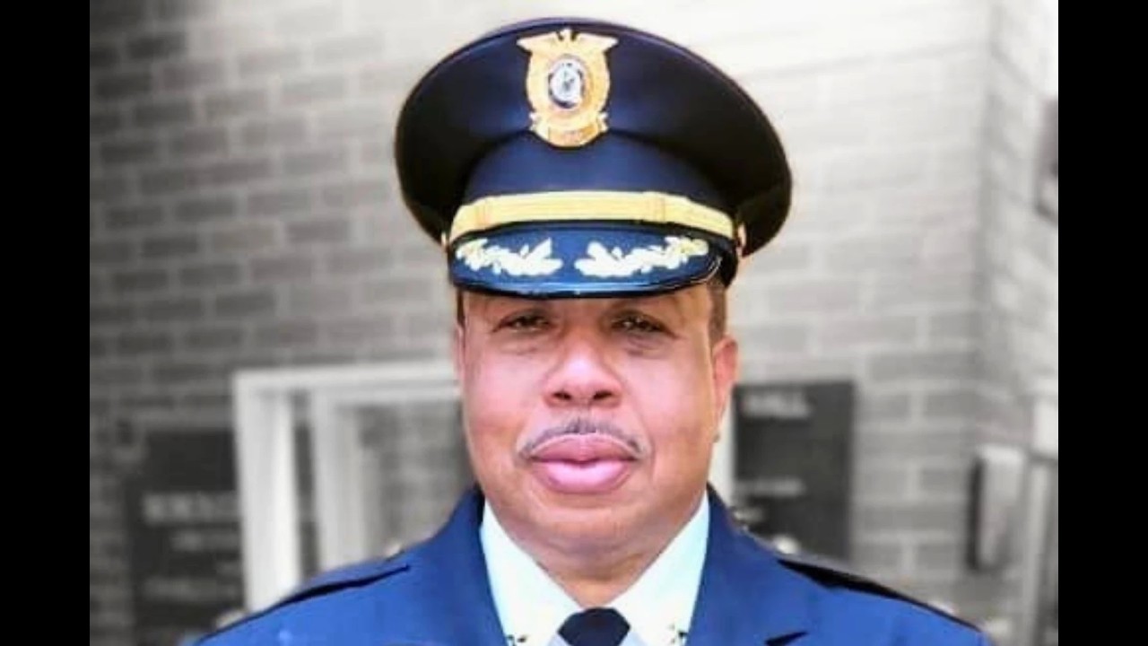 Portrait of a man in a dark blue police uniform and peaked cap with gold insignia, outdoors in front of a building.