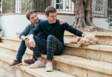 Two men sit on stone steps in a yard; one pats a small fluffy dog resting beside them.