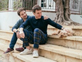 Two men sit on stone steps in a yard; one pats a small fluffy dog resting beside them.