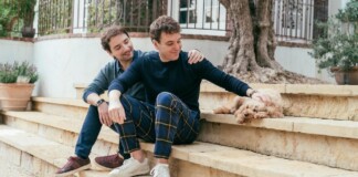 Two men sit on stone steps in a yard; one pats a small fluffy dog resting beside them.