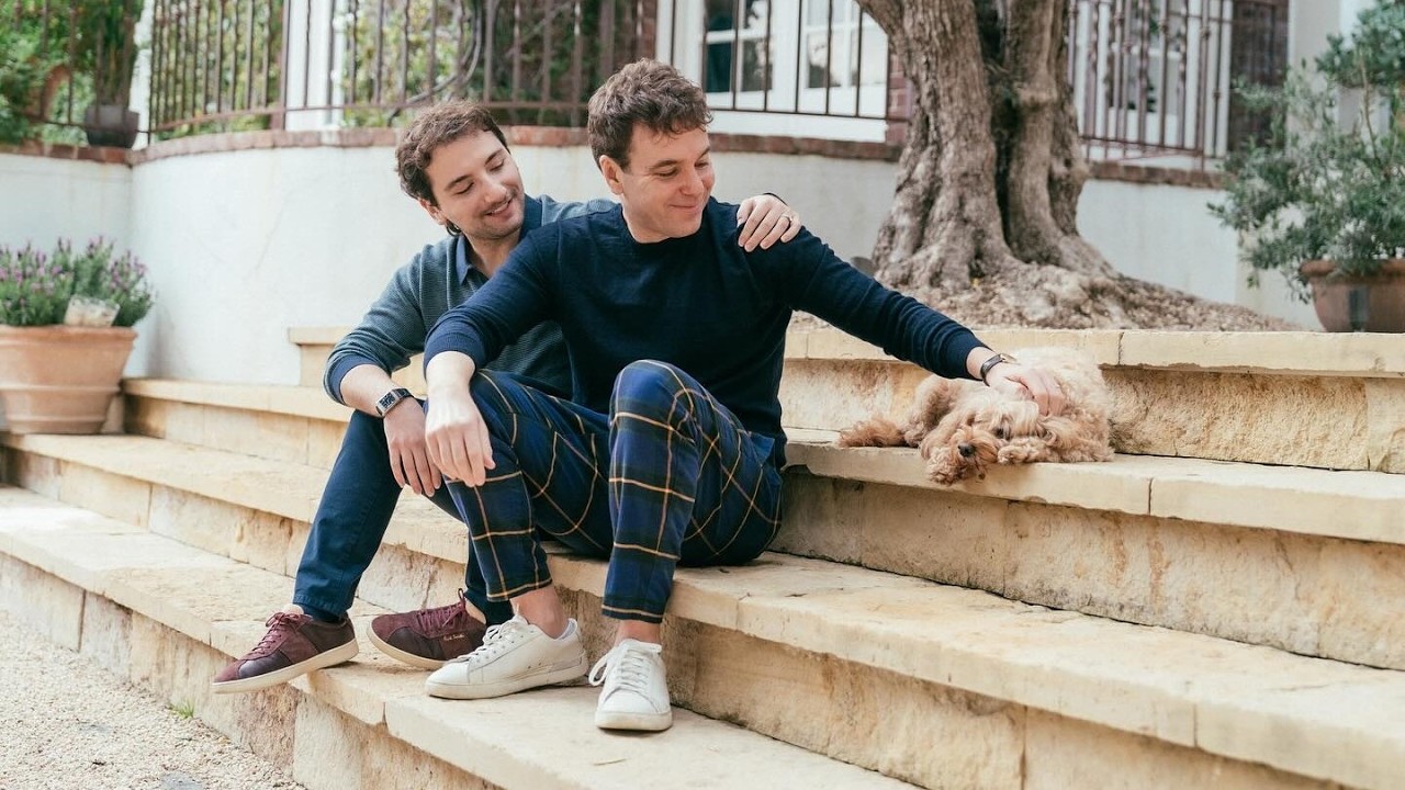 Pod Save America Host Jon Lovett Marries in Star-Studded Santa Barbara Wedding Two men sit on stone steps in a yard; one pats a small fluffy dog resting beside them.