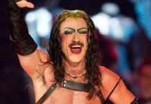 Performer with long dark hair, heavy makeup, and pearl necklace raises a gloved arm in a wrestling arena, smiling broadly.