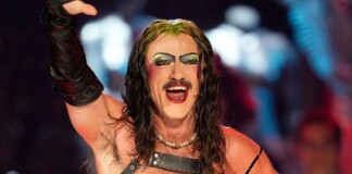 Performer with long dark hair, heavy makeup, and pearl necklace raises a gloved arm in a wrestling arena, smiling broadly.