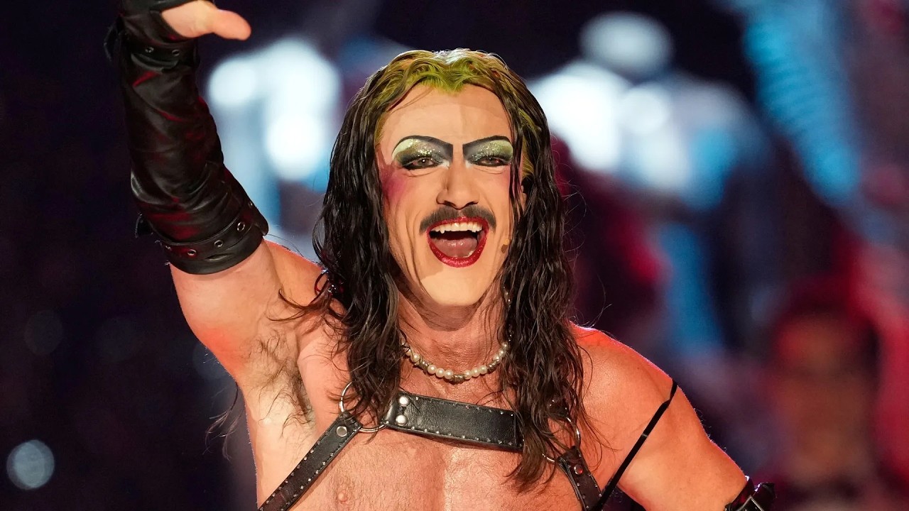 Performer with long dark hair, heavy makeup, and pearl necklace raises a gloved arm in a wrestling arena, smiling broadly.