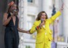 Two performers on an outdoor stage: a person in a black sleeveless outfit and a crown holding a microphone, and a second person in a bright yellow suit raising one arm while speaking into a microphone.