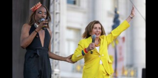 Two performers on an outdoor stage: a person in a black sleeveless outfit and a crown holding a microphone, and a second person in a bright yellow suit raising one arm while speaking into a microphone.