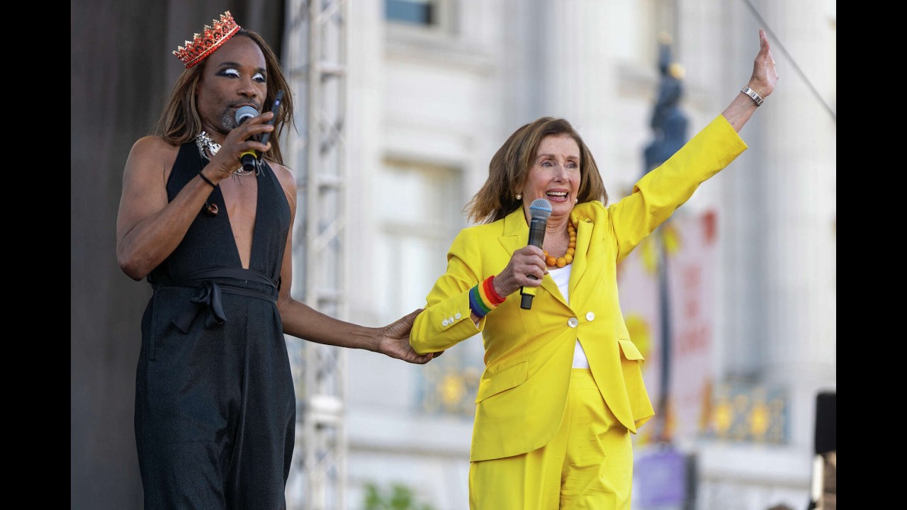 Two performers on an outdoor stage: a person in a black sleeveless outfit and a crown holding a microphone, and a second person in a bright yellow suit raising one arm while speaking into a microphone.