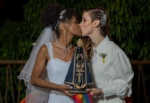 Two brides kiss while holding a beaded, ornate religious statue with a rainbow ribbon at night.