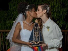 Two brides kiss while holding a beaded, ornate religious statue with a rainbow ribbon at night.