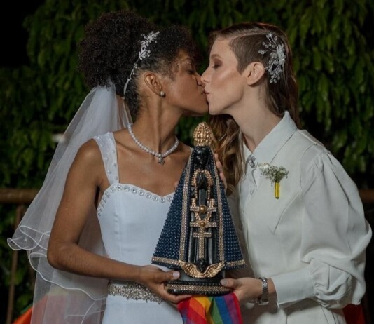 Two brides kiss while holding a beaded, ornate religious statue with a rainbow ribbon at night.