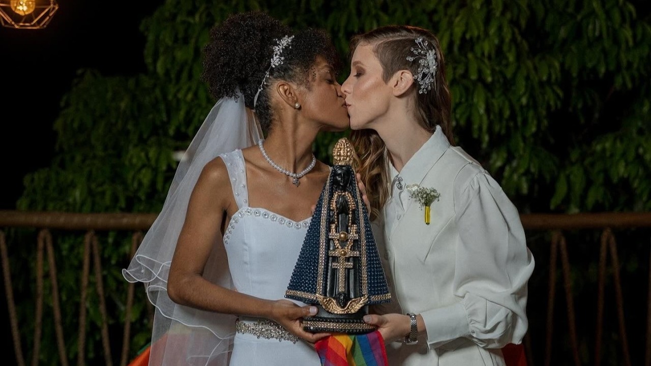 Two brides kiss while holding a beaded, ornate religious statue with a rainbow ribbon at night.