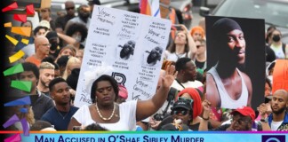 Crowd of diverse protesters holding signs and a large portrait photo, with a blue news banner at the bottom reading a hate-crime murder story.