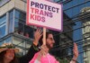 Parade participants hold a sign reading 'PROTECT TRANS KIDS' in front of a glass-fronted building.