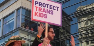 Parade participants hold a sign reading 'PROTECT TRANS KIDS' in front of a glass-fronted building.