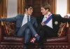 Two men in formal suits smile and lean toward each other on a red leather sofa in an elegant room, one wearing Union Jack socks and the other stars and stripes socks.