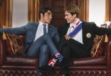 Two men in formal suits smile and lean toward each other on a red leather sofa in an elegant room, one wearing Union Jack socks and the other stars and stripes socks.