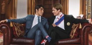 Two men in formal suits smile and lean toward each other on a red leather sofa in an elegant room, one wearing Union Jack socks and the other stars and stripes socks.