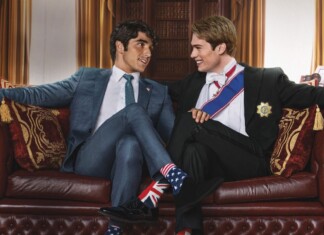 Casey McQuiston Says Red, White & Royal Wedding Sequel Will Be “Spicier” Two men in formal suits smile and lean toward each other on a red leather sofa in an elegant room, one wearing Union Jack socks and the other stars and stripes socks.