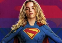 Woman in a blue Superman costume with red cape, hands on hips, against a rainbow-striped background.