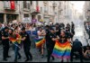Police form a barrier as LGBTQ2+ protesters march down a narrow street, holding rainbow flags and Turkish signs amid cheering crowds.