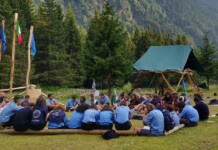 A group of scouts in blue uniforms sit in a circle on logs at a forest campsite with flag poles and tents nearby.