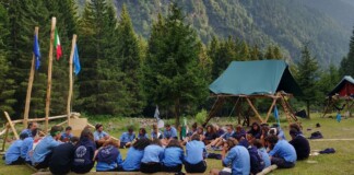 A group of scouts in blue uniforms sit in a circle on logs at a forest campsite with flag poles and tents nearby.