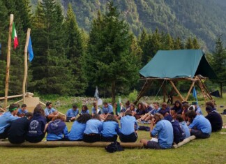 A group of scouts in blue uniforms sit in a circle on logs at a forest campsite with flag poles and tents nearby.