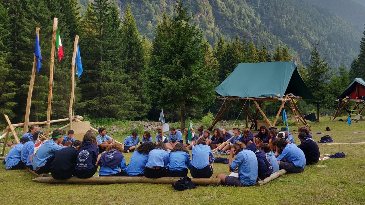 Italian Catholic Scouts Open Leadership Roles to LGBTQ+ Members for the First Time A group of scouts in blue uniforms sit in a circle on logs at a forest campsite with flag poles and tents nearby.