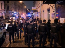 Night-time street scene with police officers and civilians gathered outside a building, a rainbow flag visible among the group, police vehicles in the background.