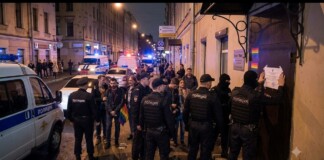 Night-time street scene with police officers and civilians gathered outside a building, a rainbow flag visible among the group, police vehicles in the background.