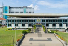 Modern government building with a wide staircase, flanked by greenery, and a central emblem above the entrance.