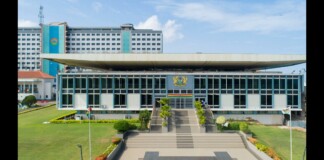 Modern government building with a wide staircase, flanked by greenery, and a central emblem above the entrance.